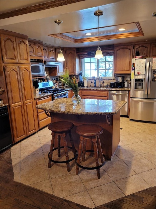 kitchen featuring light stone countertops, a tray ceiling, appliances with stainless steel finishes, under cabinet range hood, and a center island