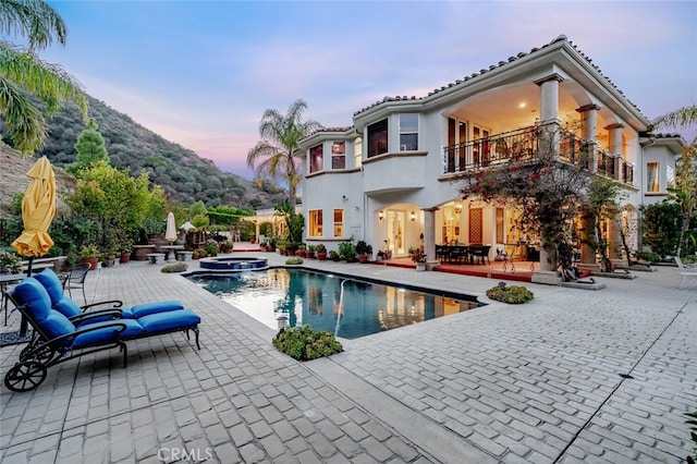 back house at dusk featuring a pool with hot tub, a balcony, and a patio area