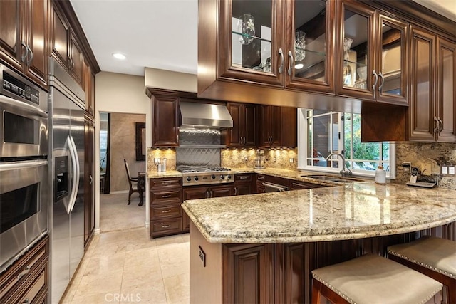 kitchen with a breakfast bar, stainless steel appliances, glass insert cabinets, wall chimney range hood, and a peninsula