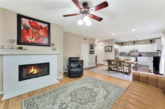 living room with a tiled fireplace, ceiling fan, light wood-type flooring, and baseboards