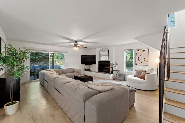 living area featuring stairs, a fireplace with raised hearth, light wood-type flooring, and a ceiling fan