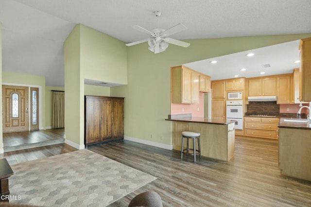 kitchen featuring light brown cabinetry, under cabinet range hood, a sink, white appliances, and a peninsula