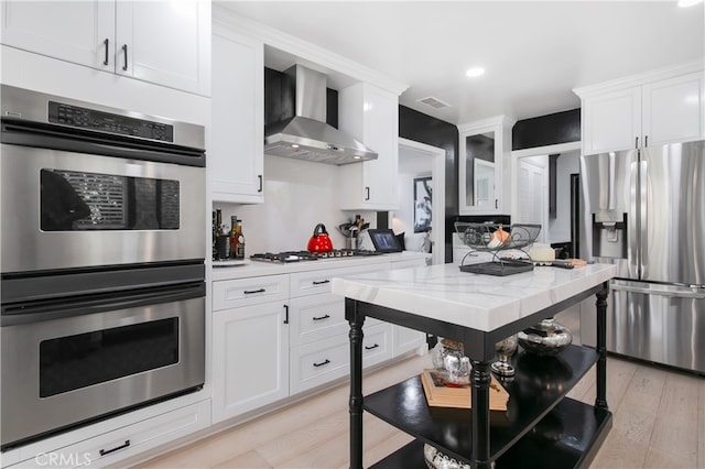 kitchen with stainless steel appliances, light stone counters, white cabinets, wall chimney range hood, and light hardwood / wood-style floors