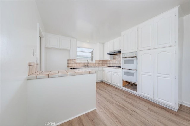 kitchen with tile countertops, decorative backsplash, double oven, white cabinetry, and under cabinet range hood