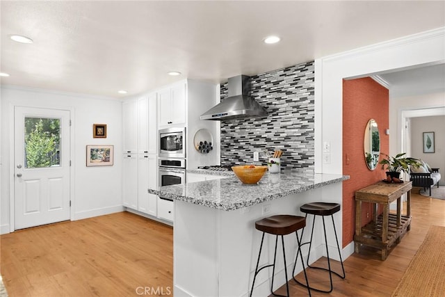 kitchen featuring wall chimney exhaust hood, stainless steel appliances, white cabinets, and light stone counters