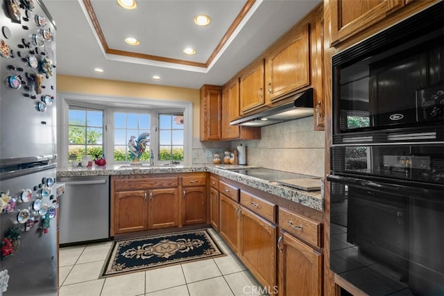 kitchen with under cabinet range hood, backsplash, black appliances, a tray ceiling, and brown cabinetry