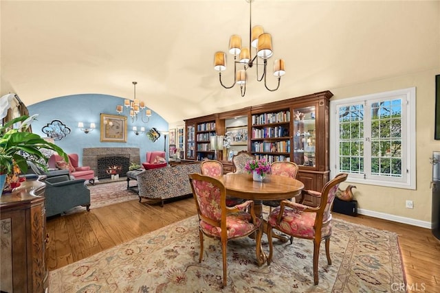 dining room featuring a notable chandelier, lofted ceiling, wood finished floors, a warm lit fireplace, and baseboards