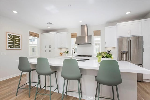 kitchen featuring white cabinets, high end fridge, light countertops, wall chimney exhaust hood, and a center island with sink