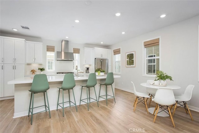 kitchen with high quality fridge, white cabinets, light countertops, wall chimney range hood, and an island with sink