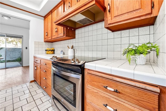 kitchen featuring premium range hood, electric range, backsplash, brown cabinetry, and tile counters