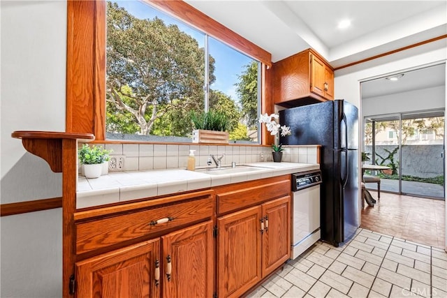 kitchen with a sink, tasteful backsplash, freestanding refrigerator, brown cabinetry, and white dishwasher