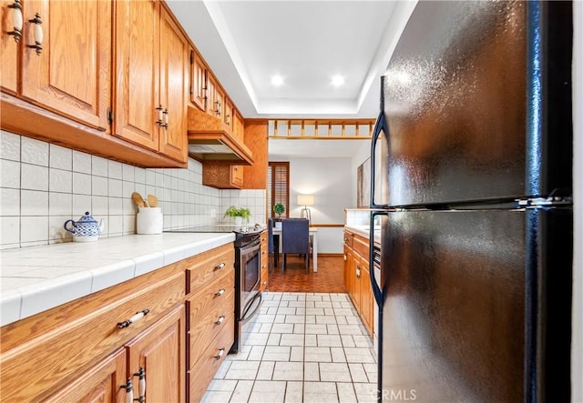 kitchen featuring tile countertops, brown cabinetry, freestanding refrigerator, stainless steel range with electric stovetop, and backsplash