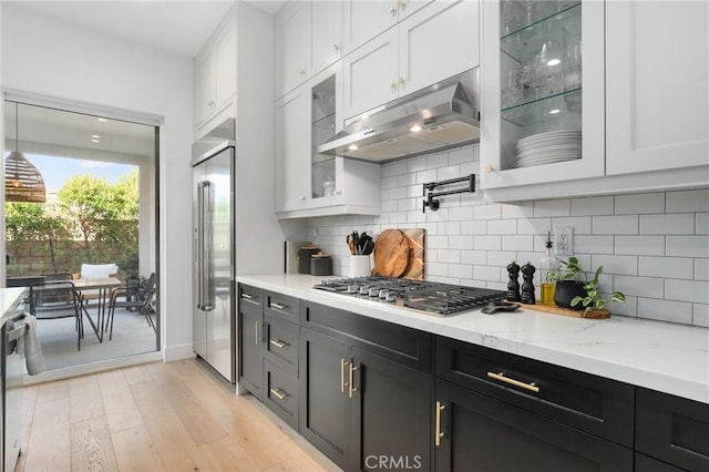 kitchen with stainless steel appliances, glass insert cabinets, white cabinets, light stone countertops, and under cabinet range hood