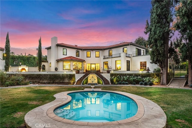 back house at dusk featuring a lawn, a fenced in pool, and a balcony