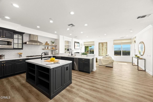 kitchen with an island with sink, wall chimney range hood, stainless steel microwave, ornamental molding, and sink