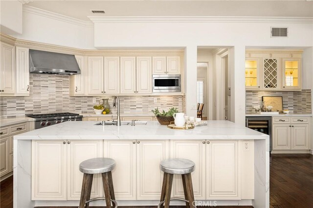 kitchen with a breakfast bar area, light stone countertops, wall chimney exhaust hood, stainless steel microwave, and a center island with sink