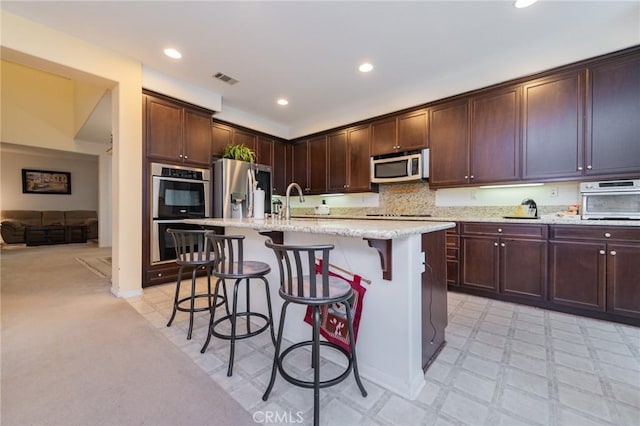 kitchen featuring a center island with sink, dark brown cabinets, light stone counters, appliances with stainless steel finishes, and a kitchen breakfast bar