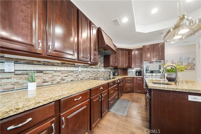 kitchen with light wood finished floors, custom range hood, light stone countertops, black electric cooktop, and a sink