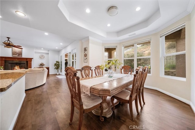 dining space featuring dark wood finished floors, recessed lighting, a raised ceiling, a lit fireplace, and baseboards