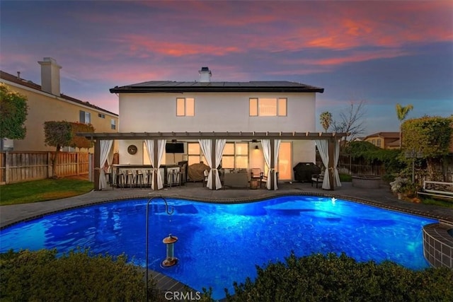 back of property at dusk with outdoor dry bar, stucco siding, solar panels, a patio area, and a fenced backyard