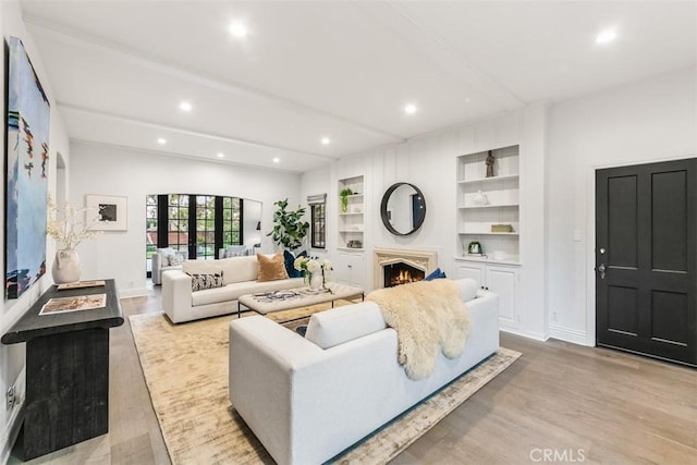 living room with a warm lit fireplace, light wood-style flooring, beamed ceiling, built in shelves, and recessed lighting