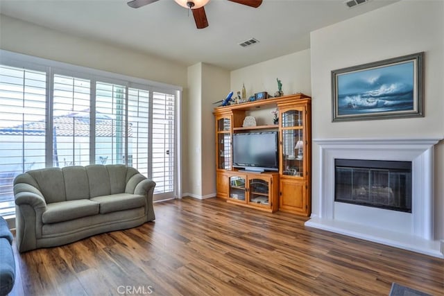 living area featuring dark wood-style floors, ceiling fan, visible vents, and a glass covered fireplace