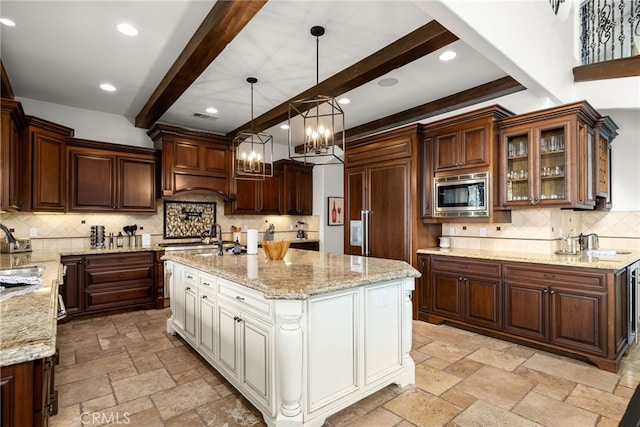 kitchen with stainless steel microwave, custom range hood, hanging light fixtures, an island with sink, and light stone countertops