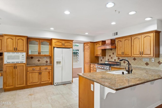 kitchen featuring a peninsula, white appliances, a sink, wall chimney exhaust hood, and glass insert cabinets