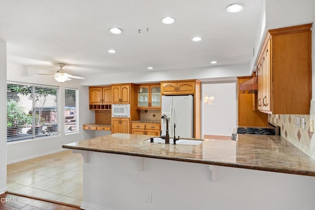 kitchen featuring brown cabinetry, glass insert cabinets, light stone countertops, white appliances, and a peninsula