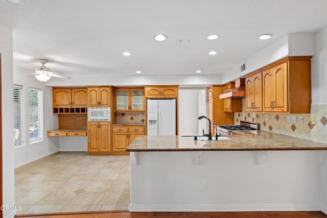 kitchen with a peninsula, white appliances, brown cabinetry, and wall chimney range hood