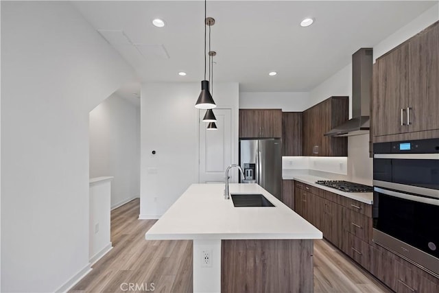 kitchen featuring an island with sink, hanging light fixtures, stainless steel appliances, sink, and wall chimney range hood