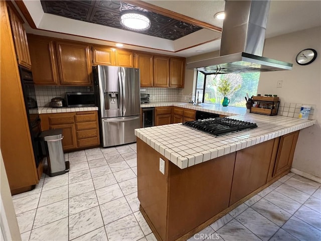 kitchen featuring tile countertops, black appliances, island exhaust hood, a raised ceiling, and kitchen peninsula