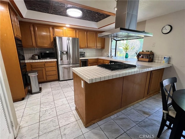 kitchen featuring appliances with stainless steel finishes, tile counters, island exhaust hood, a raised ceiling, and kitchen peninsula