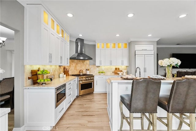 kitchen with crown molding, light wood-style flooring, wall chimney range hood, built in appliances, and a kitchen breakfast bar