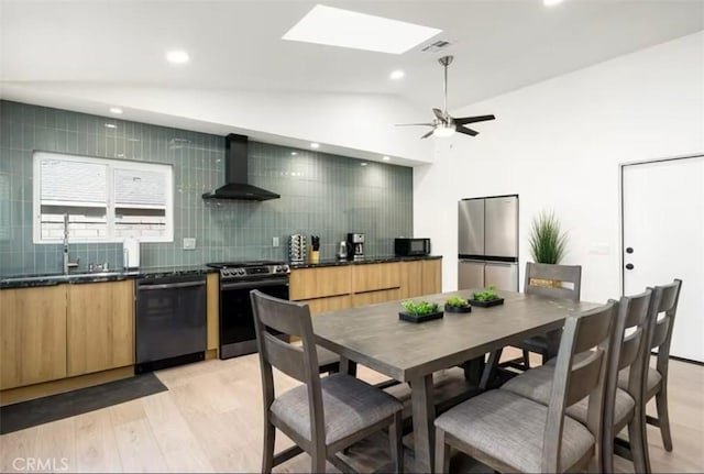 kitchen with vaulted ceiling with skylight, wall chimney exhaust hood, black appliances, light hardwood / wood-style floors, and backsplash