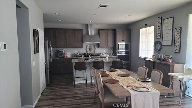 kitchen featuring appliances with stainless steel finishes, an island with sink, light stone counters, wall chimney range hood, and a breakfast bar