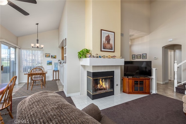 tiled living area with arched walkways, a tiled fireplace, baseboards, ceiling fan with notable chandelier, and high vaulted ceiling