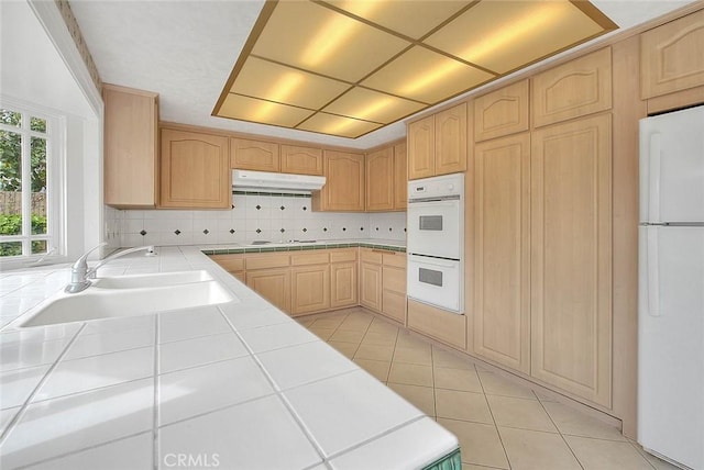 kitchen with white appliances, tile countertops, a sink, light brown cabinetry, and under cabinet range hood