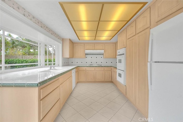 kitchen with light brown cabinetry, a sink, under cabinet range hood, tile countertops, and white appliances