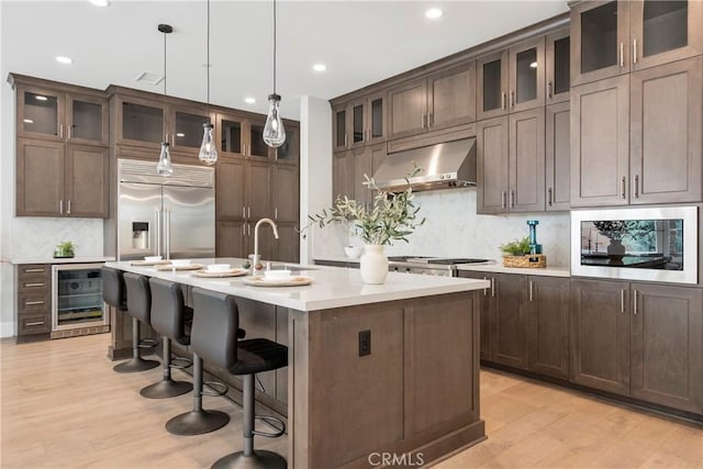 kitchen with built in appliances, decorative light fixtures, beverage cooler, light wood-type flooring, and a kitchen island with sink