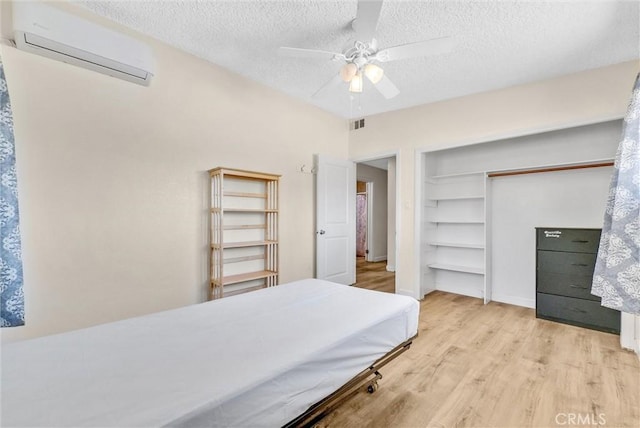 bedroom featuring an AC wall unit, light hardwood / wood-style flooring, a closet, ceiling fan, and a textured ceiling