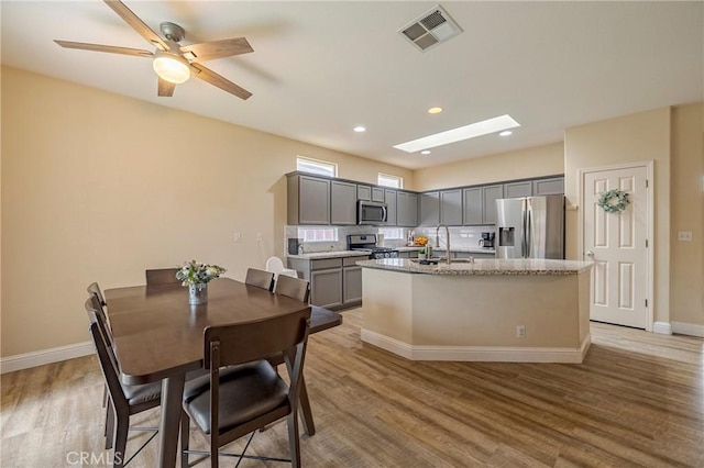 kitchen featuring a center island with sink, gray cabinetry, stainless steel appliances, light stone countertops, and sink