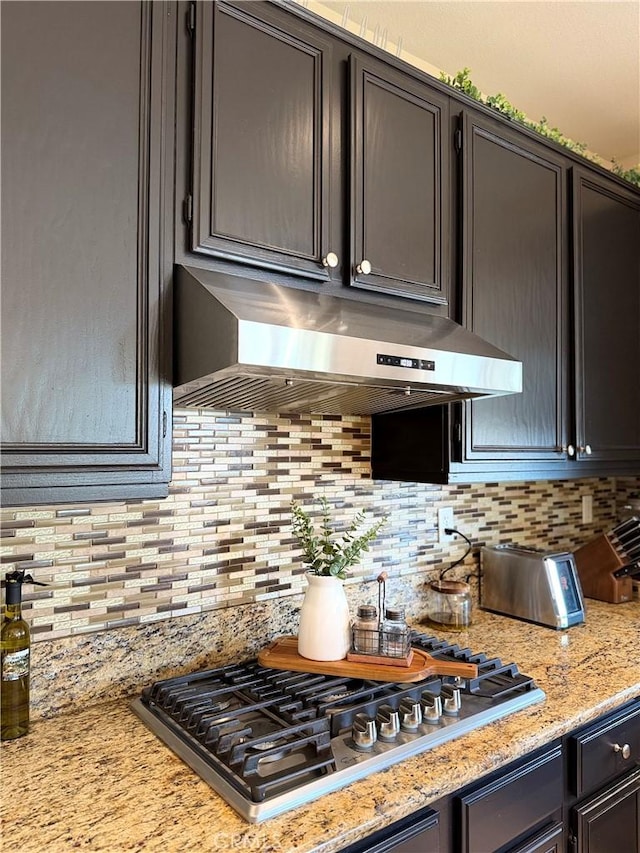 kitchen featuring light stone counters, backsplash, stainless steel gas stovetop, and under cabinet range hood