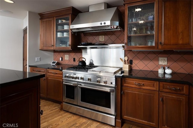 kitchen featuring range with two ovens, light wood-type flooring, wall chimney exhaust hood, dark countertops, and glass insert cabinets