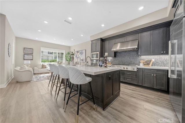 kitchen with a center island with sink, backsplash, built in appliances, under cabinet range hood, and a kitchen bar