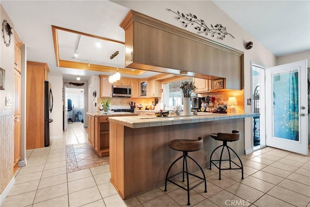 kitchen with a kitchen bar, stainless steel microwave, glass insert cabinets, a peninsula, and tile countertops