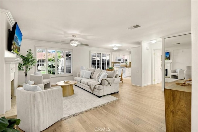 living room with visible vents, a ceiling fan, crown molding, a fireplace, and light wood-style floors