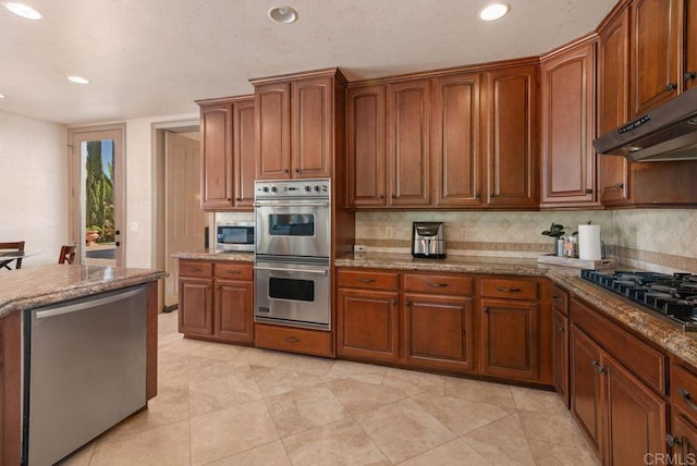 kitchen featuring under cabinet range hood, appliances with stainless steel finishes, brown cabinets, backsplash, and light stone countertops