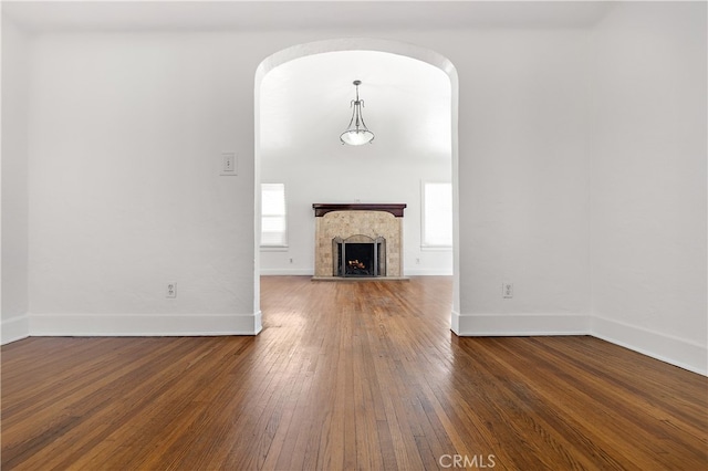 unfurnished living room featuring hardwood / wood-style floors, arched walkways, baseboards, and a warm lit fireplace