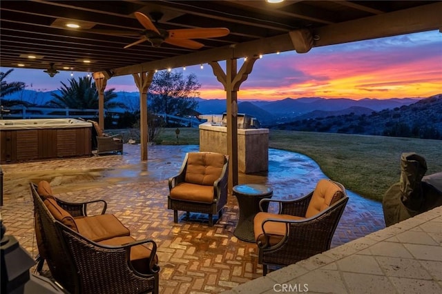 patio terrace at dusk with a mountain view, a hot tub, and ceiling fan